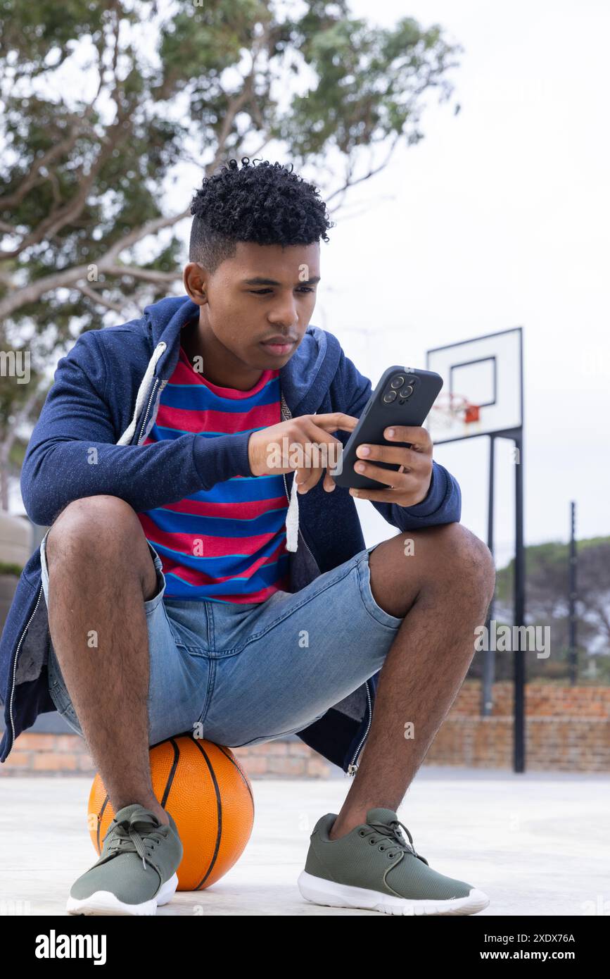 Sitzt auf Basketball, Teenager mit Smartphone auf dem Platz im Freien, konzentriert sich intensiv Stockfoto