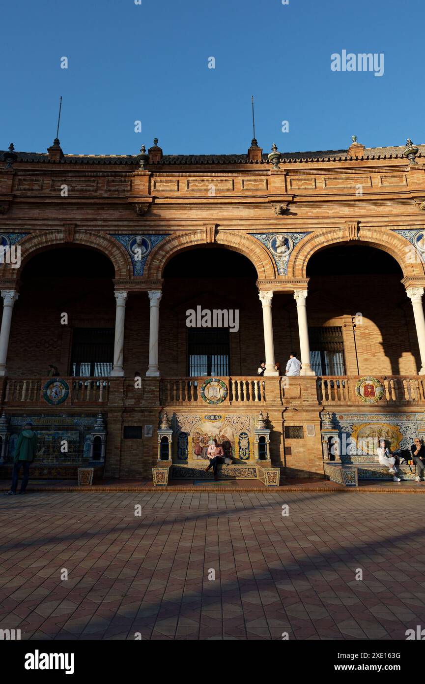 Besucher, der auf einer kunstvoll gekachelten Bank für Coruña an der Plaza de España in Sevilla sitzt, umgeben von komplexen architektonischen Details und sonnendurchfluteten Bögen Stockfoto