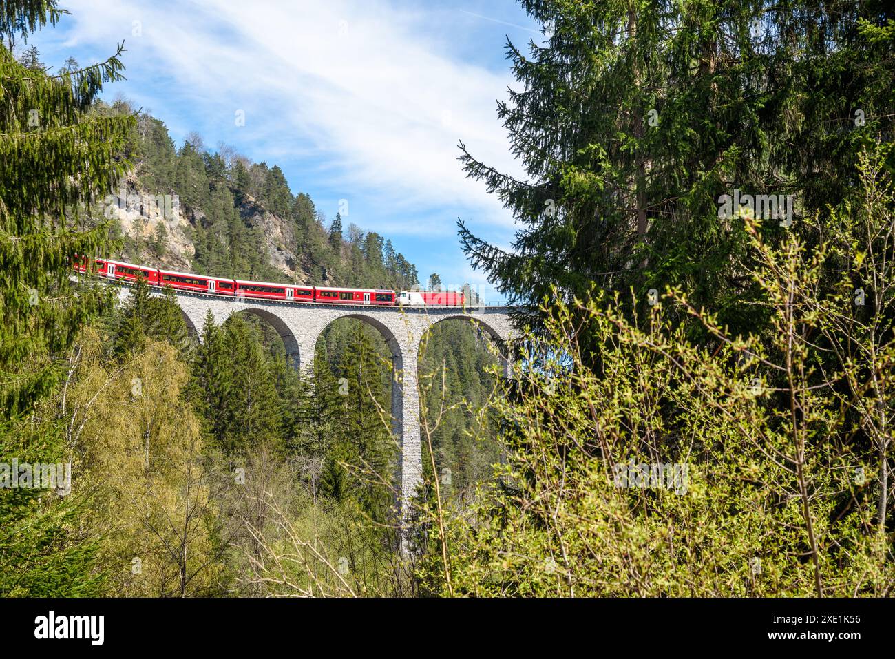 Roter Personenzug über einem hohen Steinviadukt in den Bergen an einem sonnigen Frühlingstag Stockfoto