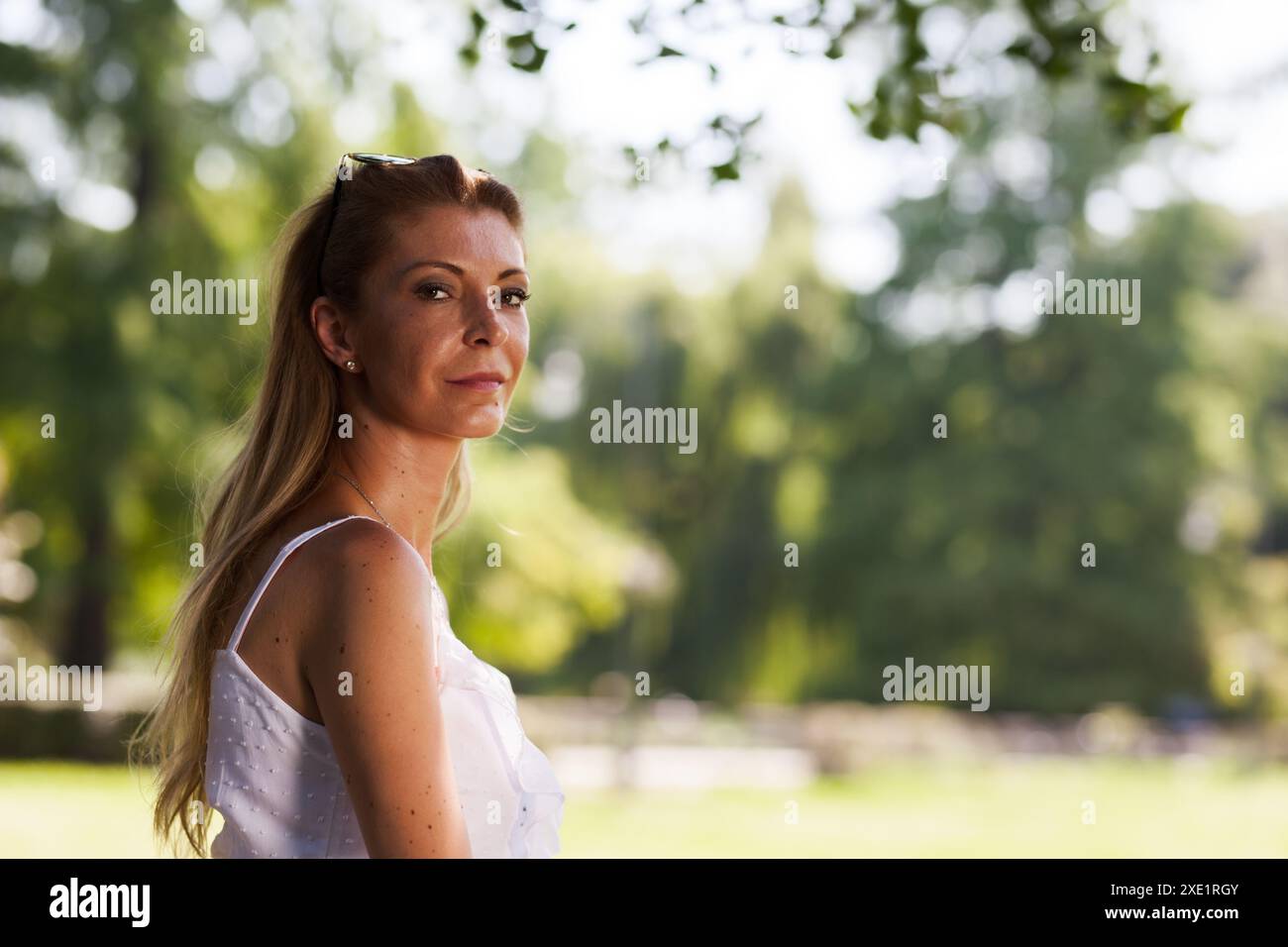 Eine Frau mittleren Alters in einem weißen ärmellosen Kleid steht selbstbewusst in einem üppigen Park, wo das Sonnenlicht durch die Bäume gefiltert wird. Stockfoto