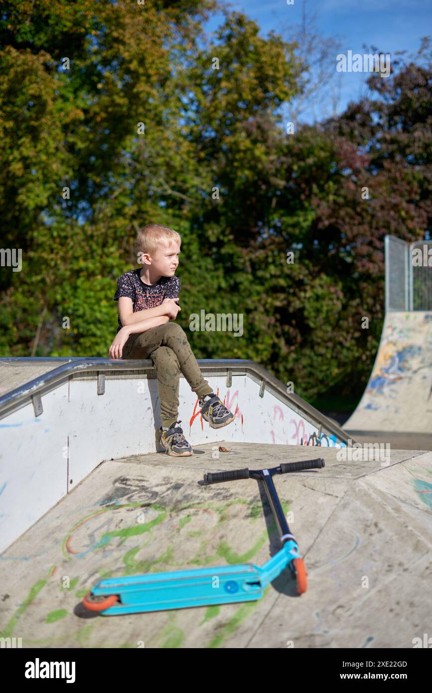 Junge auf dem Roller macht einen Trick und genießt seine Fahrt im Skatepark am bewölkten Frühlingstag. Junger Mann, der auf dem Kick sitzt Stockfoto