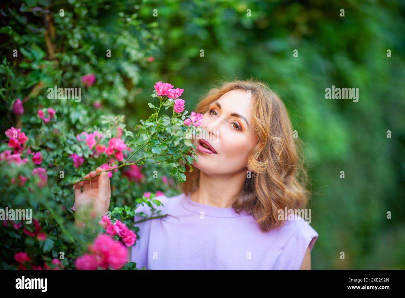 Schöne erfolgreiche Frau mittleren Alters, die blühende Rosen riecht und im Garten draußen genießt. Stockfoto