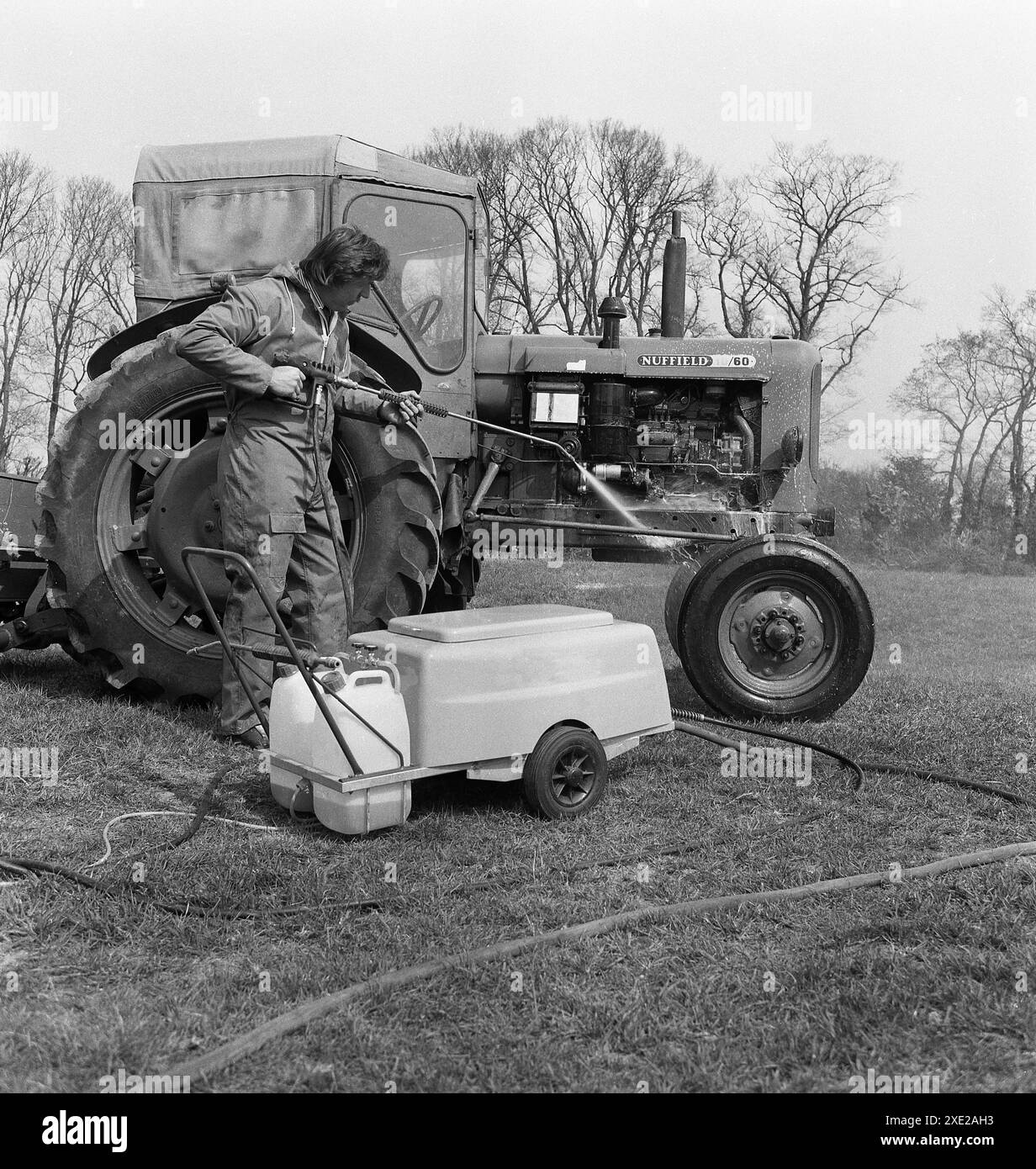 1970er Jahre, historisch, draußen auf einem Feld, ein Mann, der mit einem elektrischen Strahlreiniger einen Nuffield 10/60 Dieseltraktor mit Schutzhaube reinigt, England, Großbritannien. Der von BMC hergestellte Traktor wurde 1964 eingeführt und war der erste der Nuffield-Traktoren, die serienmäßig mit Power-Tag-off und hydraulischem Gestänge ausgestattet waren. Stockfoto