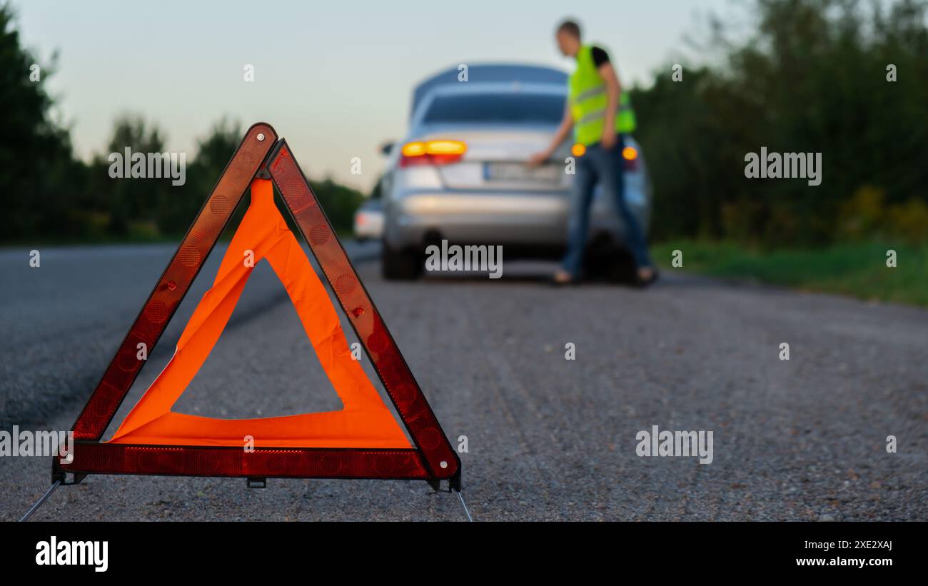 Gebrochenes Silber Luxusauto Notfall Unfall. Man-Fahrer mit rotem Dreieck-Stoppschild auf der Straße. Sportauto eingeschaltet bl Stockfoto