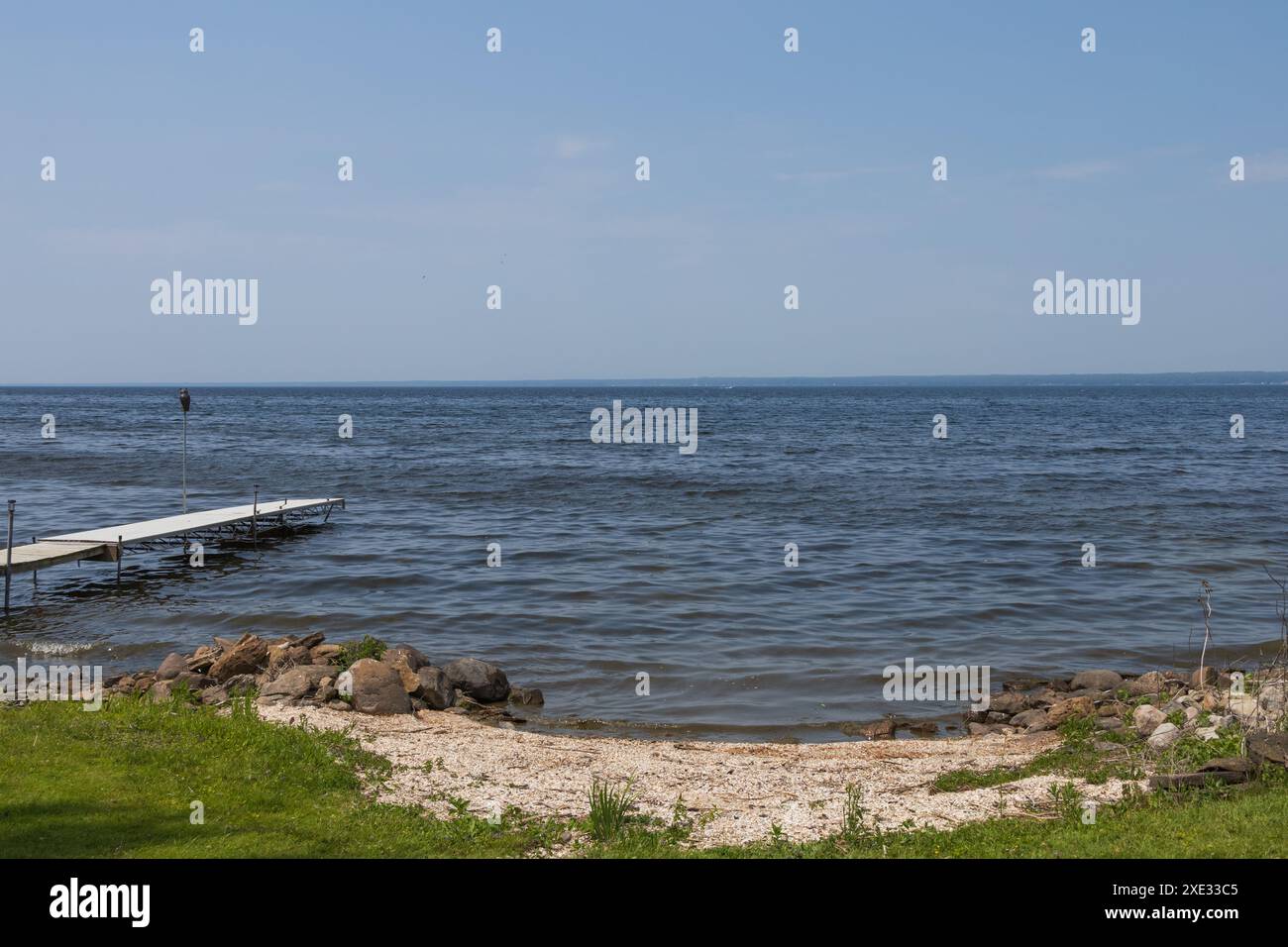 Blick auf einen See und Horizont mit einem kleinen Sandstrand und einem Bootssteg. Keine Menschen oder Boote. An einem sonnigen, klaren Tag. Stockfoto