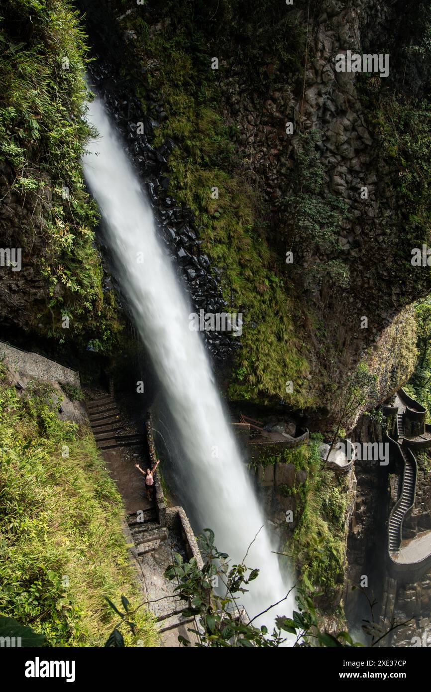 Die vertikale junge Latina steht mit erhobenen Armen unter dem majestätischen Wasserfall Pailon del Diablo und seiner gewundenen Steintreppe in Baños de Agua San Stockfoto