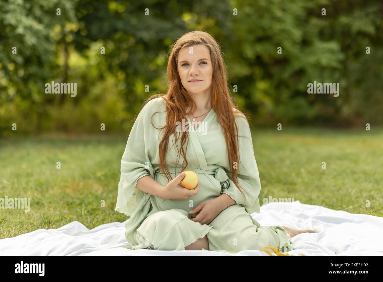 Weiße, hübsche schwangere Frau sitzt auf einer Decke, hält in der Hand und isst Apfel im Park. Wiese genießen, Entspannung. Gesunder Lebensstil. Die Entbindung Stockfoto