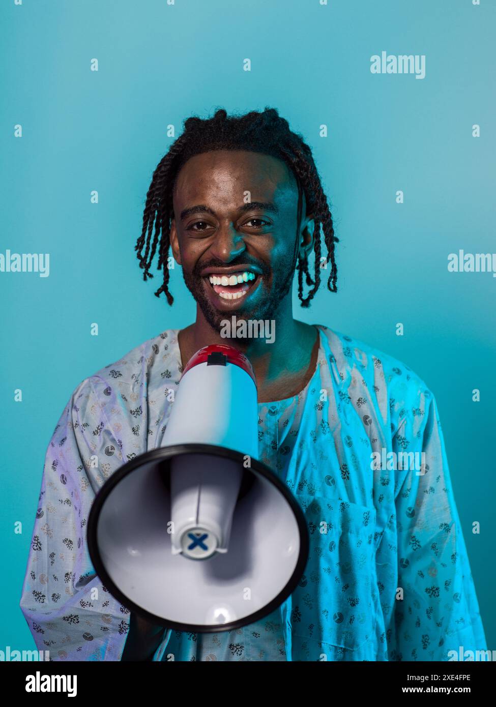 Der afroamerikanische Mann trägt traditionelle Kleidung und verwendet leidenschaftlich ein Megaphon vor einem auffälligen blauen Hintergrund, was symbolisiert Stockfoto