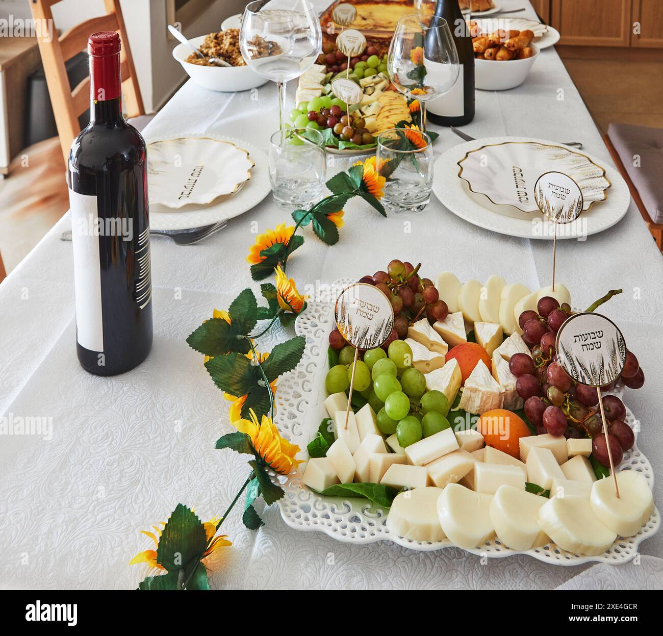 Ein eleganter Shavuot-Feiertagstisch mit Wein, Käse, Obst und Weihnachtsdekoration, bereit für ein traditionelles jüdisches Feiertagsessen. Happy Shavuot Inscript Stockfoto