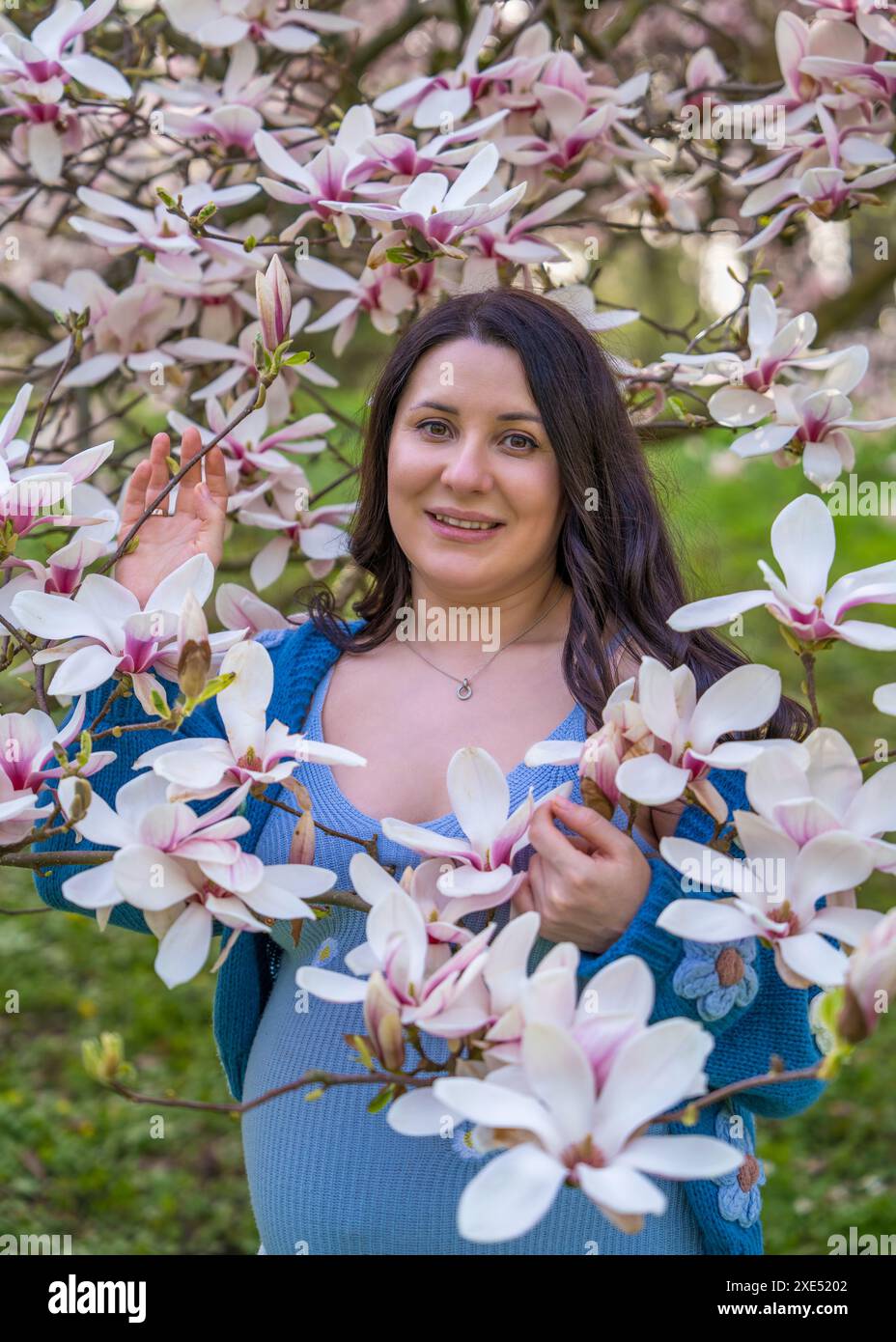 Porträt einer niedlichen schwangeren Frau im Park, gekleidet in einem blauen Kleid zwischen Magnoli-Blumen. Lady, die in die Kamera schaut, Nahaufnahme. Schwangerschaft. Warten auf Stockfoto