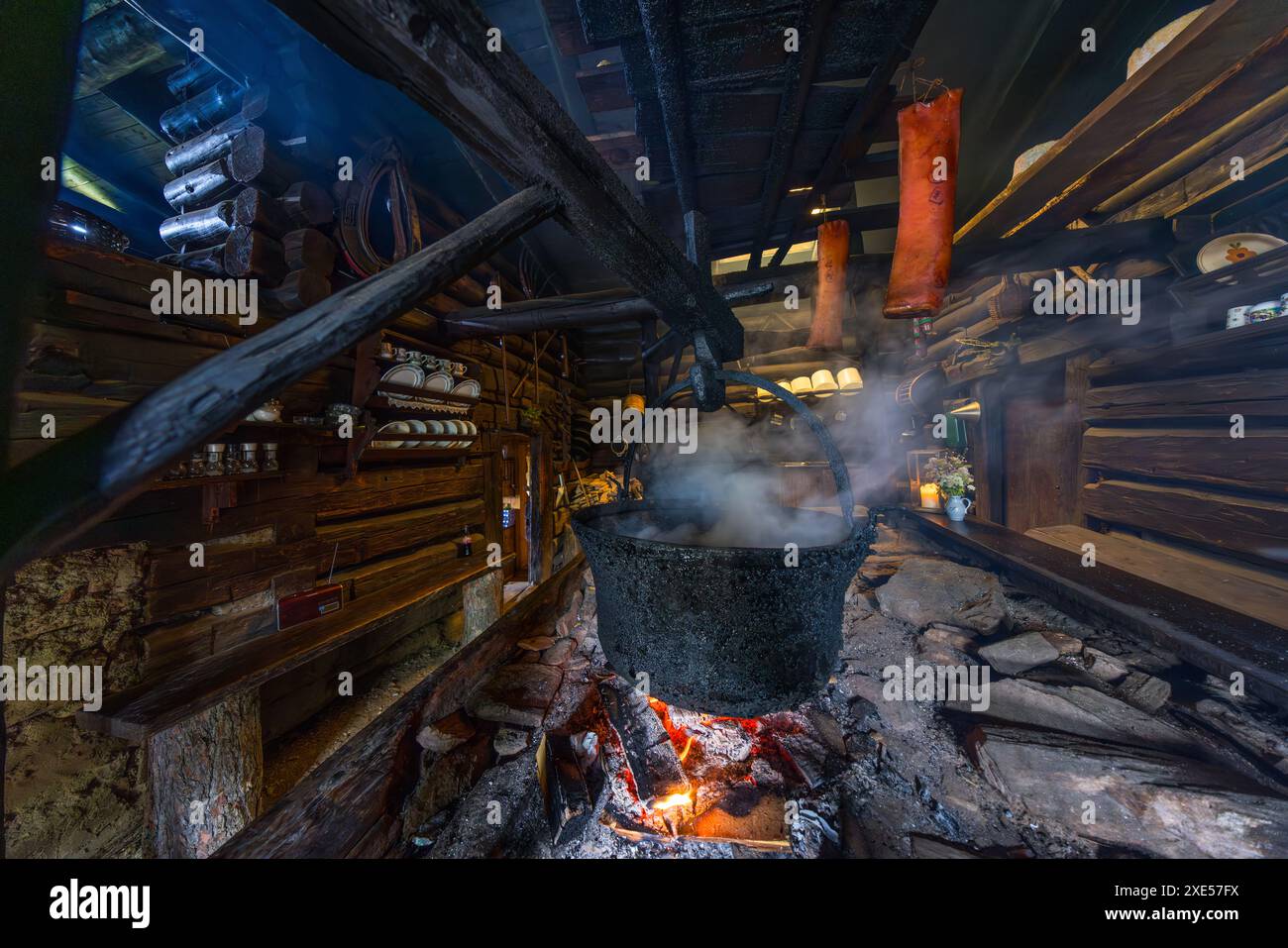 Käseblasen in einem großen Topf über dem offenen Feuer auf der Karseggalm im Großartal, während Speckschalen daneben zum Räuchern hängen, Salzburg, Österreich. Der Kamin in der 400 Jahre alten Almhütte wird noch heute zum Käse machen und zum Räuchern von Speck genutzt. Stockfoto