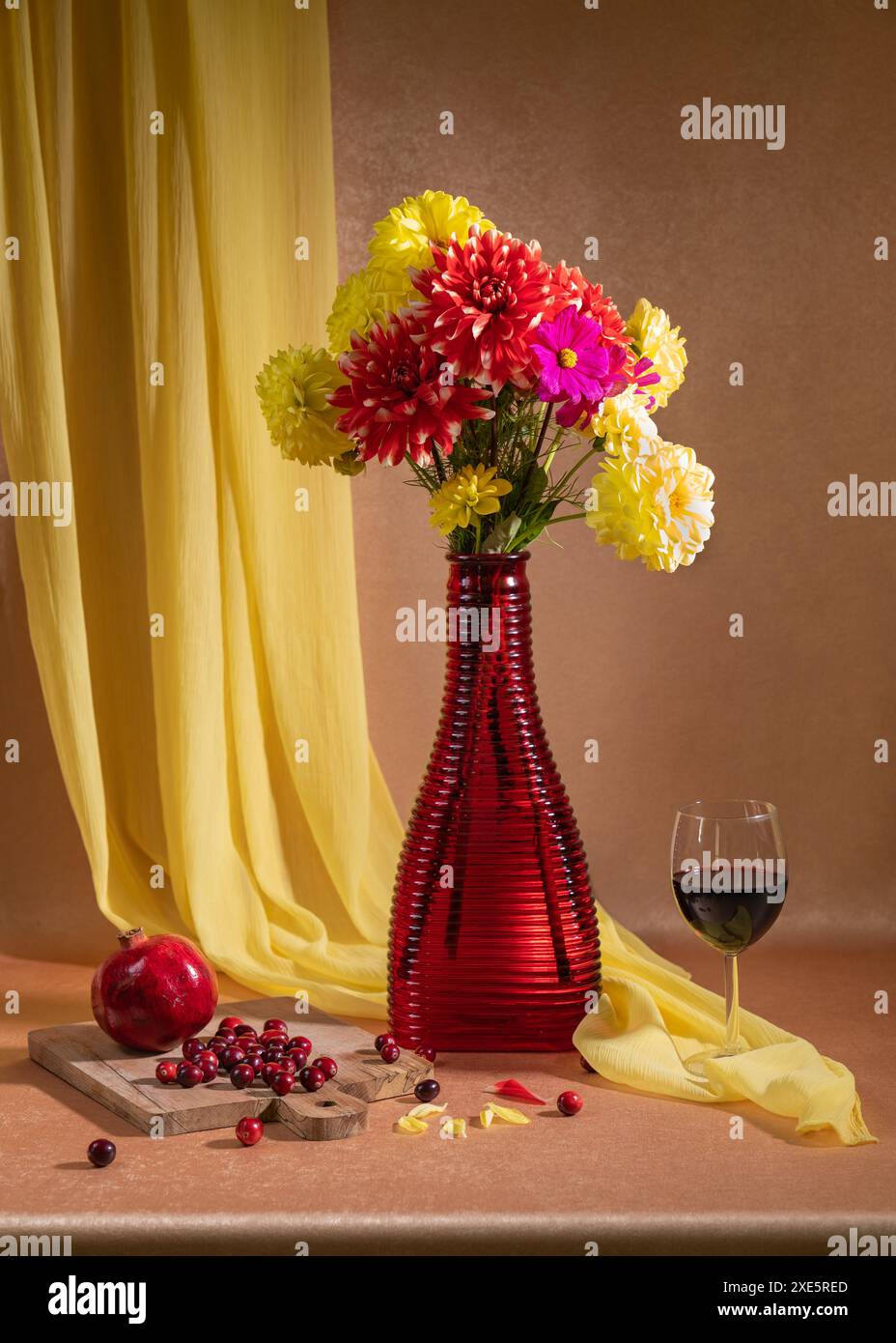 Es gibt viele helle Blumen in einer roten Vase, ein Glas Rotwein steht in der Nähe, rote Beeren und Granatäpfel liegen. Orangefarbener Hintergrund und gelber Handker Stockfoto