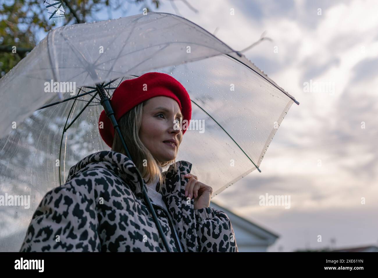Eine Frau, die einen roten Hut und einen Mantel mit Leopardenmuster trägt, hält einen klaren Regenschirm. Der Schirm ist offen und die Frau blickt in den Himmel. Die Stockfoto