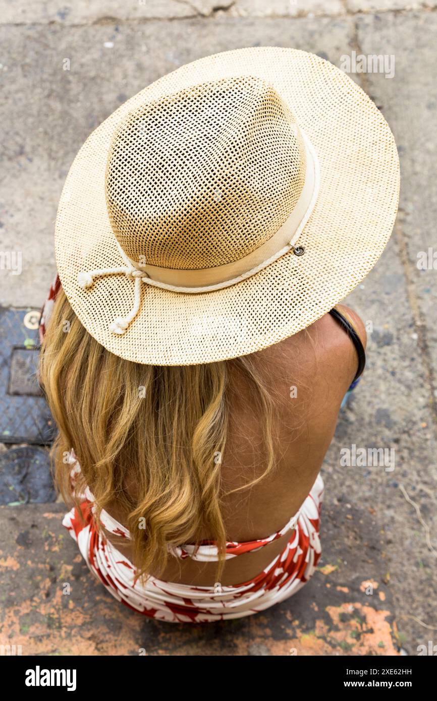 Blick von oben auf eine Frau mit hellem Strohhut und Blumenkleid, die auf einem Bürgersteig der Stadt sitzt. Ideal für Themen wie Sommer, Mode und urbanes Leben Stockfoto