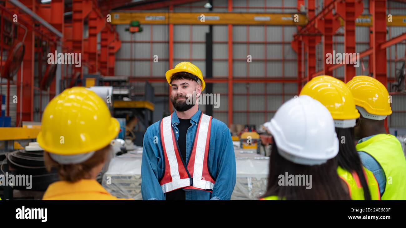 Vorgesetzter und Gruppe von Fabrikarbeitern, die Schutzhelme tragen, treffen sich kurz zusammen, bevor sie mit der Arbeit beginnen. Stockfoto