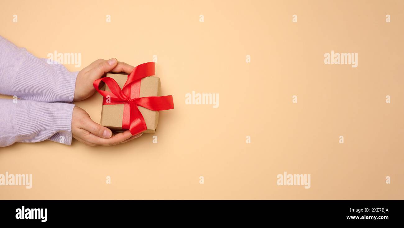 Die Hand einer Frau hält eine Geschenkbox, die in ein rotes Seidenband auf beigefarbenem Hintergrund gewickelt ist, von oben Stockfoto