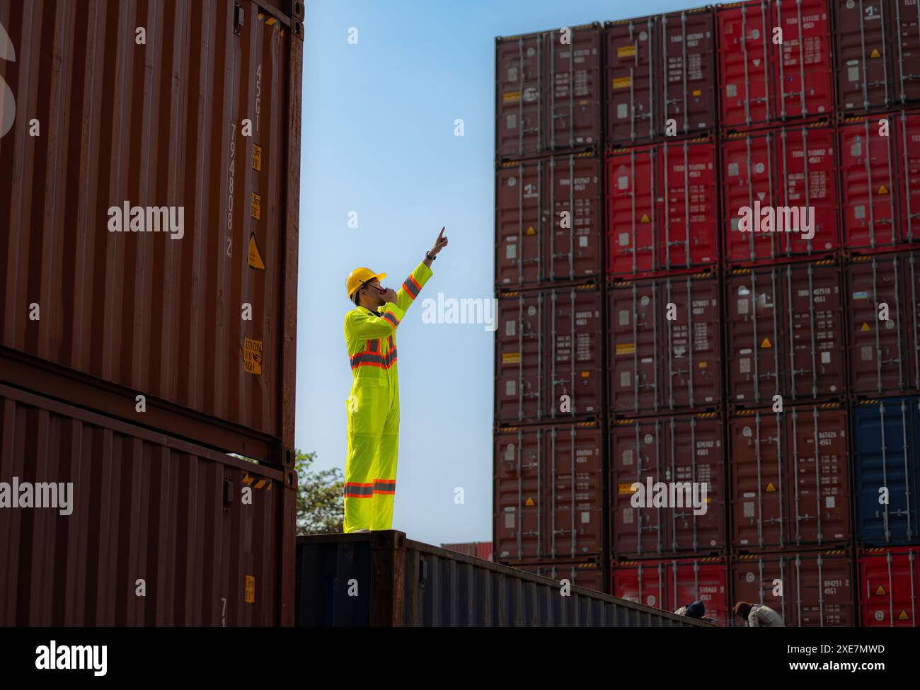 Ein männlicher Arbeiter steht auf einer Containerbox, während er einen Sicherheitsanzug trägt. Verwenden Sie das Walkie-Talkie, um mit Kollegen auf dem zu kommunizieren Stockfoto