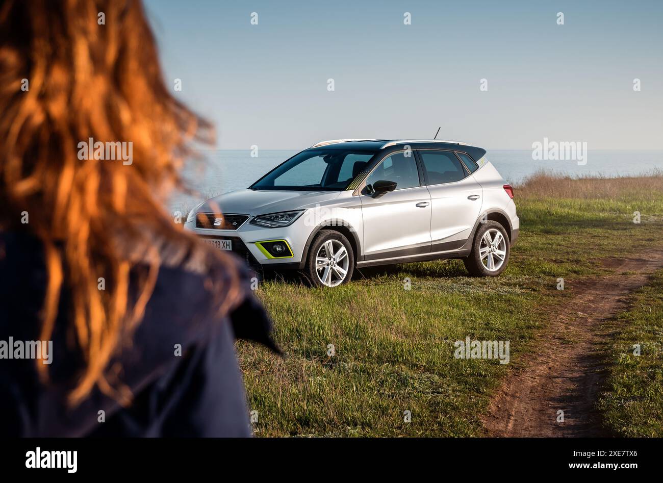 Frau, die sich einem silbernen SITZ nähert, Arona parkte auf Gras auf einer Seite einer unbefestigten Straße - über die Schulter. Drei Viertel Vorderansicht des spanischen Fließhecks. Stockfoto