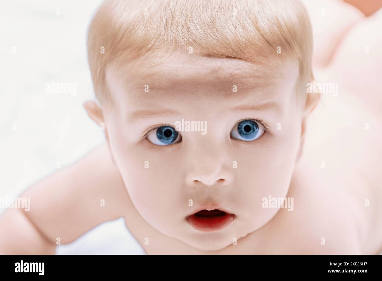 Der Junge mit blauen Augen und blonden Haaren liegt auf einer weißen Decke und blickt mit neugierigem und unschuldigem Ausdruck überraschend auf Stockfoto