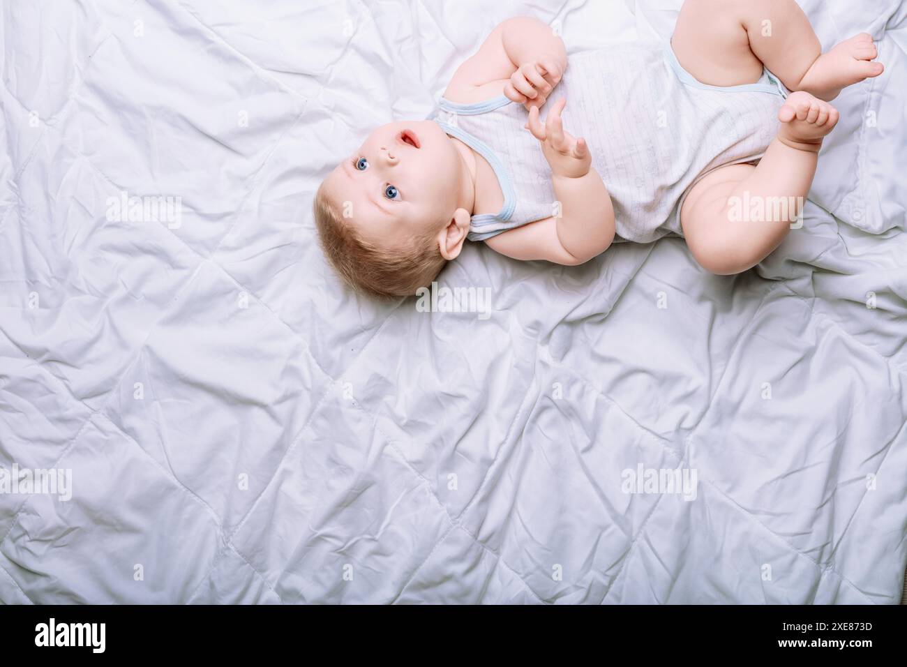 Friedliches Baby mit fesselnden blauen Augen liegt auf einem weißen Bett in einem sonnendurchfluteten Raum und strahlt Unschuld und Glück aus. Die Draufsicht bietet Kopierraum für des Stockfoto