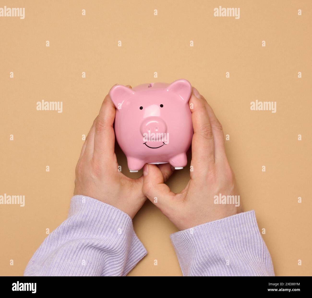 Weibliche Hände halten eine rosafarbene Keramik-Schweinebank vor braunem Hintergrund Stockfoto