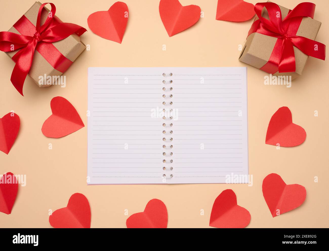 Offenes Notizbuch mit leeren weißen Blättern, roten Herzen und Geschenkkartons auf beigefarbenem Hintergrund Stockfoto