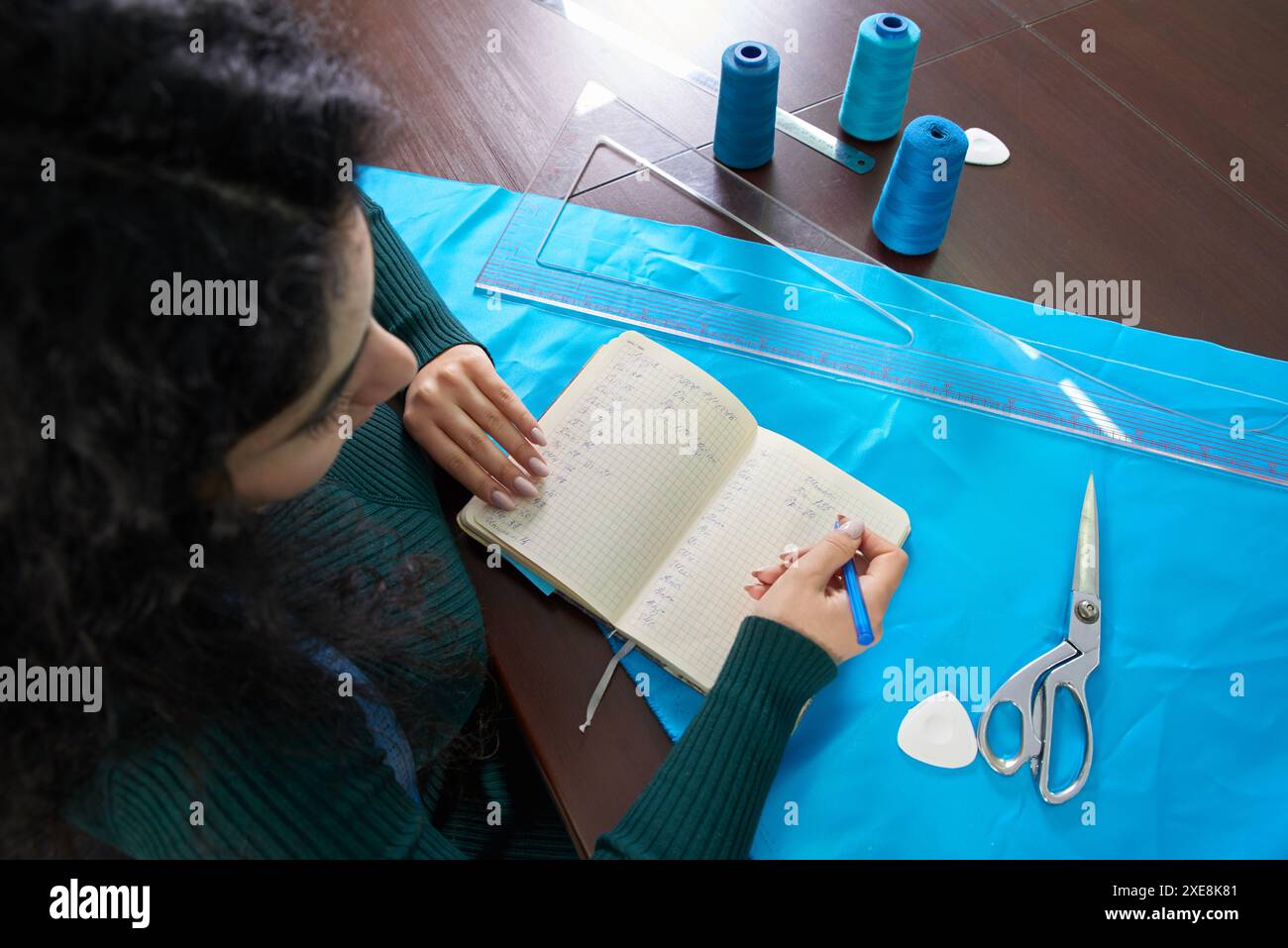 Näherin schreibt Kennzahlen in ein Notizbuch. Der Schneider kreiert eine Kollektion von Outfits. Eine junge Designerin macht mir Notizen von Ideen Stockfoto
