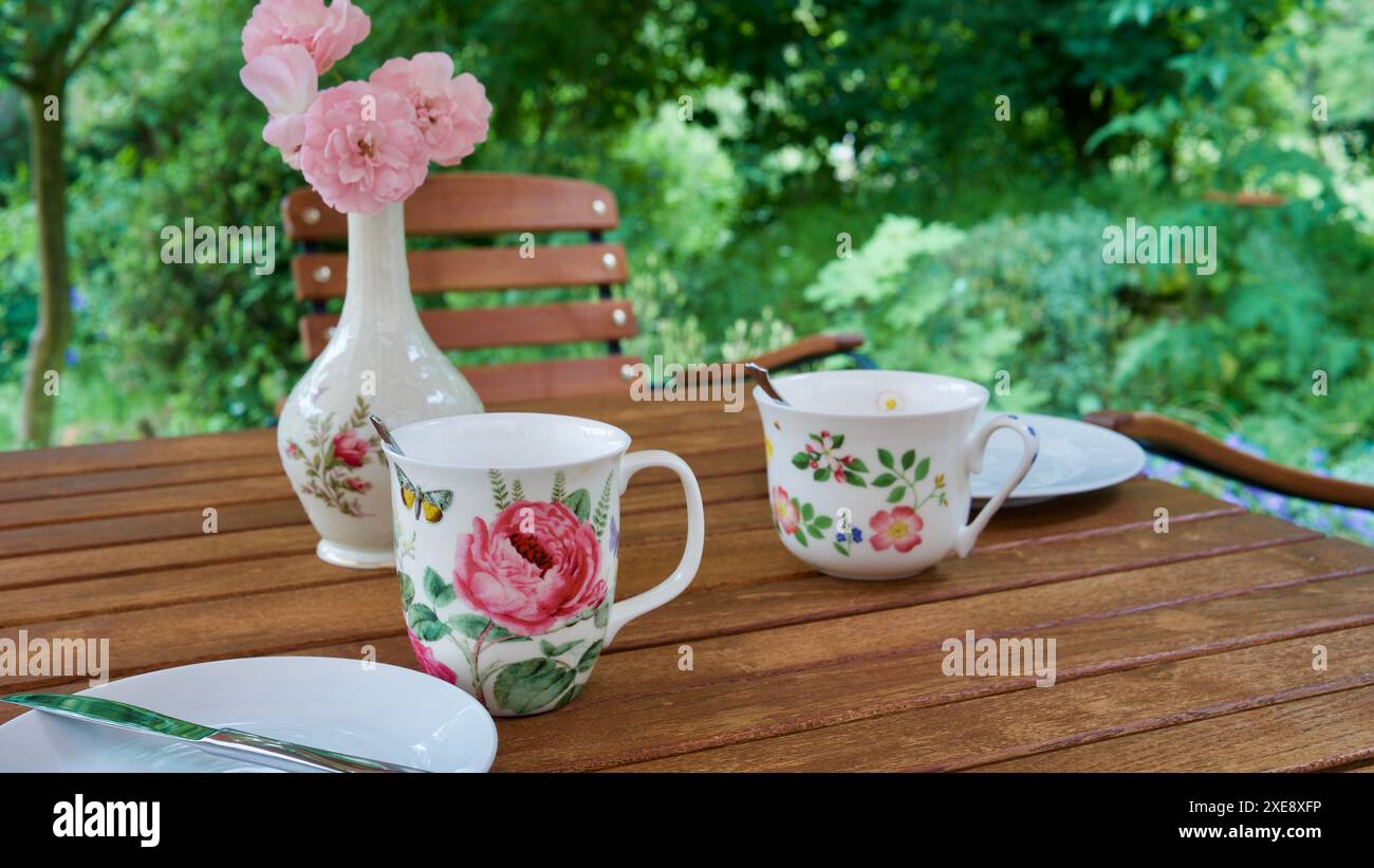 Gedeckter Tisch mit zwei hübschen Tassen, Tellern, einer Vase mit einer Rose auf einem hölzernen Gartentisch. Stockfoto