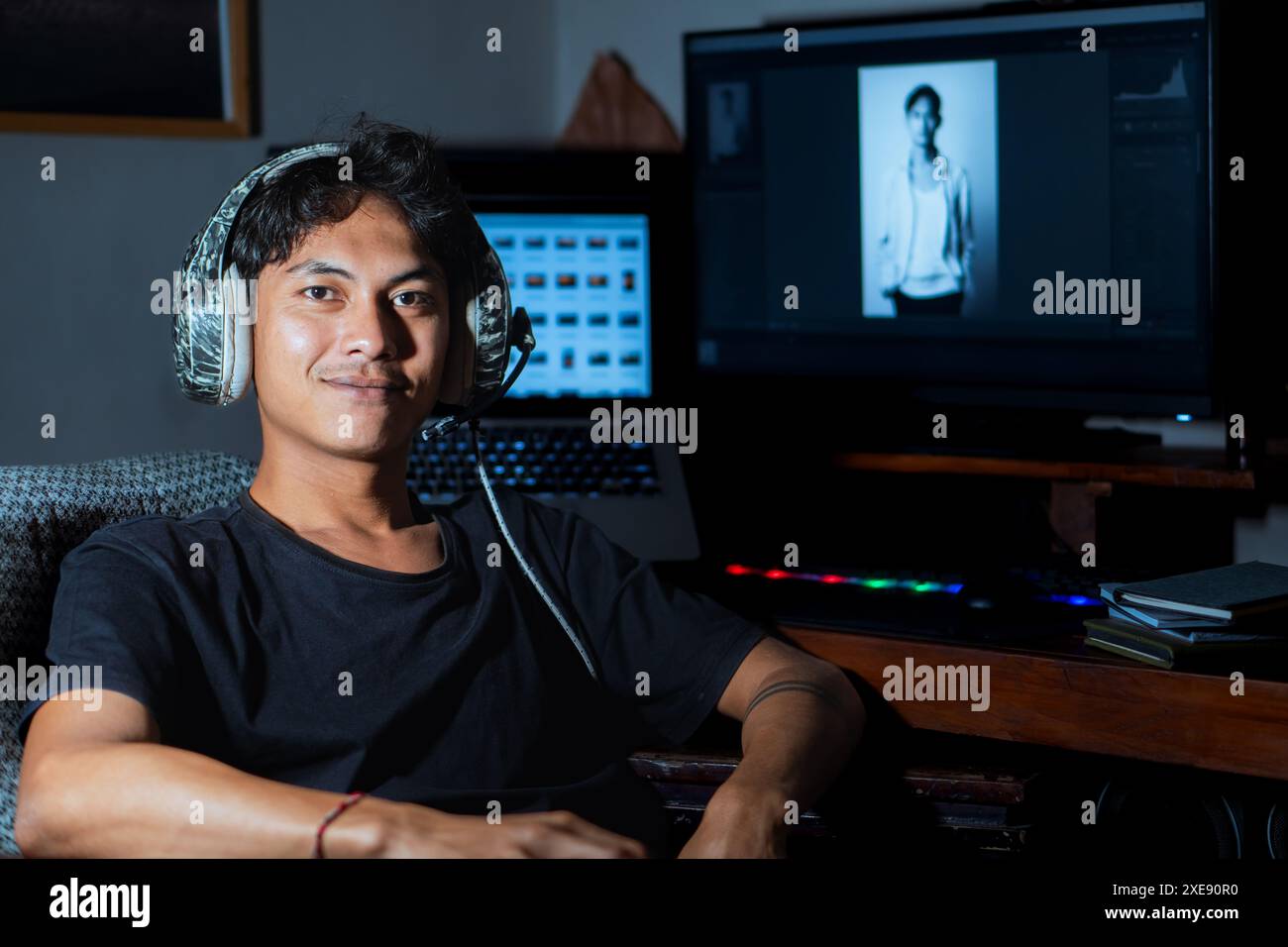 Ein junger Mann sitzt an einem Schreibtisch mit einem Computer und Kopfhörern um den Hals und lächelt in die Kamera. Auf dem Computerbildschirm wird ein ph-wert in Schwarzweiß angezeigt Stockfoto