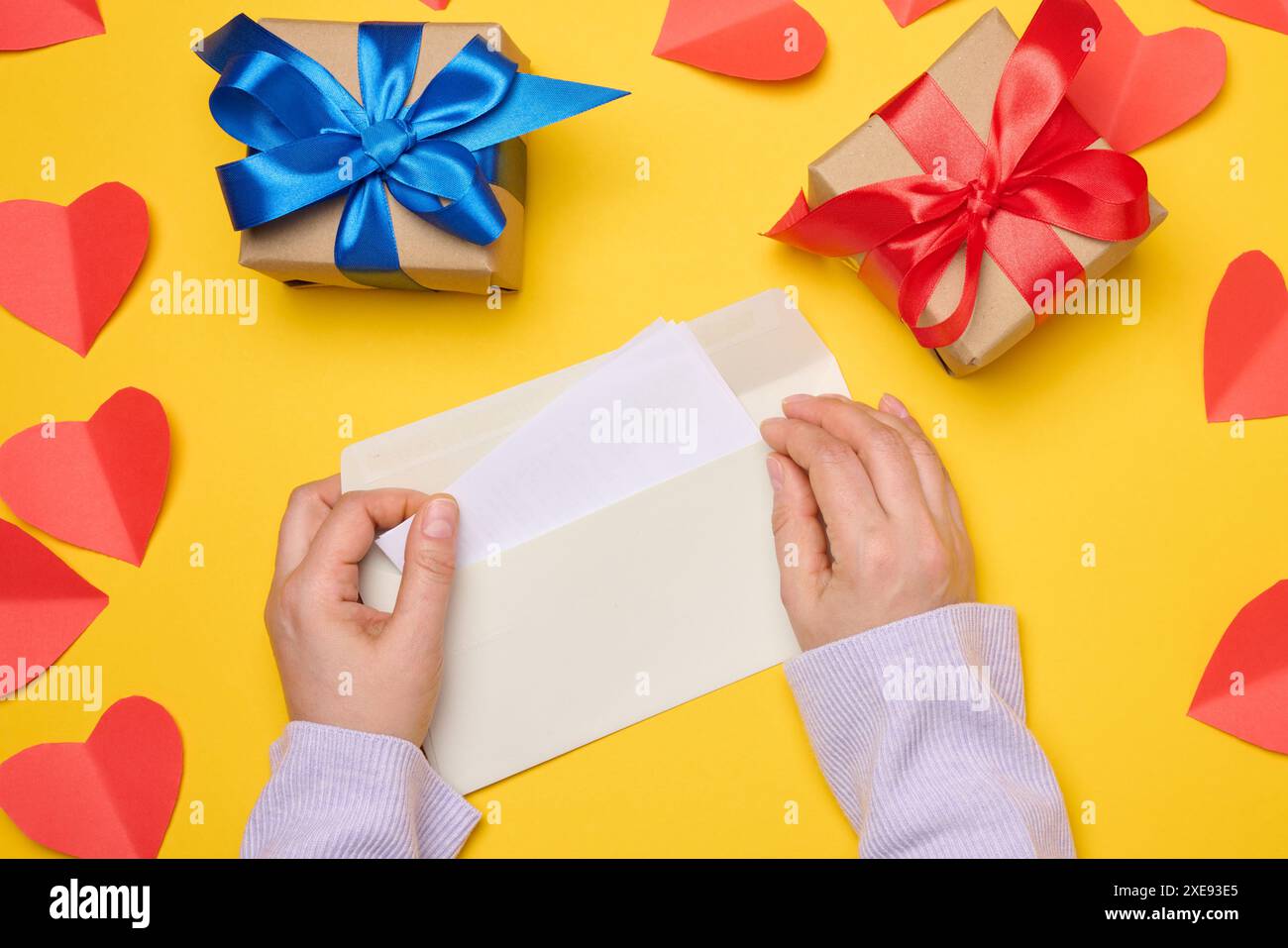 Weibliche Hände halten einen weißen rechteckigen Umschlag auf gelbem Hintergrund, papierrote Herzen. V Stockfoto