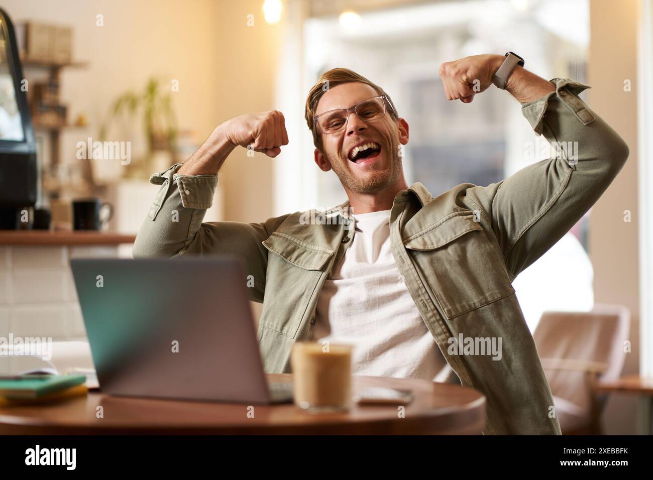 Porträt des gutaussehenden, zufriedenen jungen Mannes zeigt Bizeps, beugt Muskeln, sitzt vor dem Laptop im Café, freut sich, privater Trainer Stockfoto