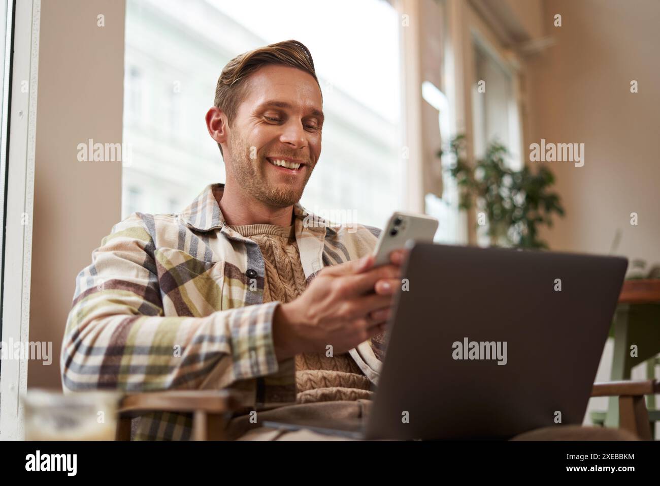 Nahaufnahme Porträt eines hübschen lächelnden Mannes mit Laptop, Café-Besucher, der auf Stuhl sitzt, Smartphone benutzt, glücklich aussieht, Nachrichten sendet Stockfoto