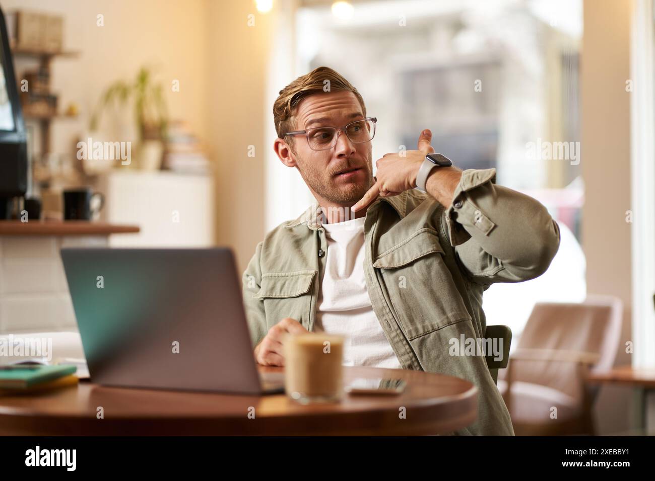 Porträt eines jungen gutaussehenden Mannes in Brille, Geschäftsmann, der im Café arbeitet, mit Laptop sitzt, Handgeste für Telefonanrufe zeigt, Anzeige Stockfoto