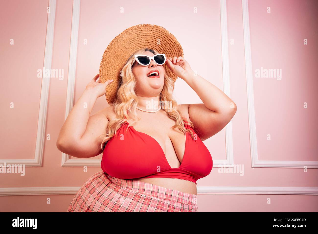 Glückliche Frauen in Übergröße, die Sommeroutfits tragen, stehen vor rosa Hintergrund, während sie die Sonnenbrille berühren und nach oben schauen Stockfoto