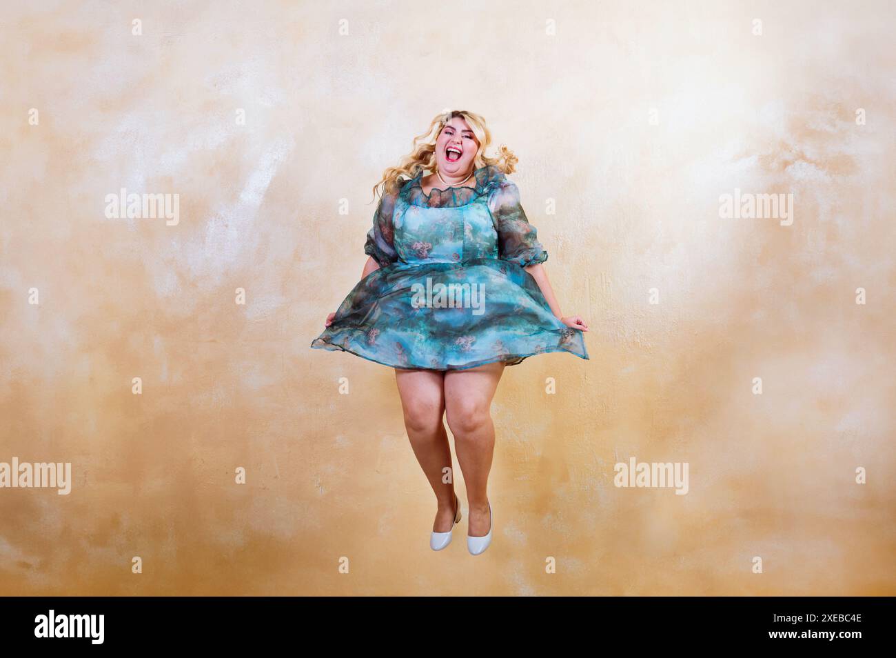 Eine Frau in Übergröße mit einem fröhlichen Ausdruck wird mitten im Sprung gefangen, ihr Kleid fließend, vor einem warmen Studio-Hintergrund Stockfoto