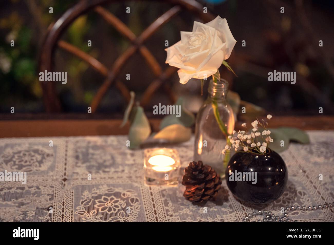 Weiße Rosenblüte in Glasflasche mit Kerzenlicht, Kiefernzapfen auf hölzernem Esstisch Stockfoto