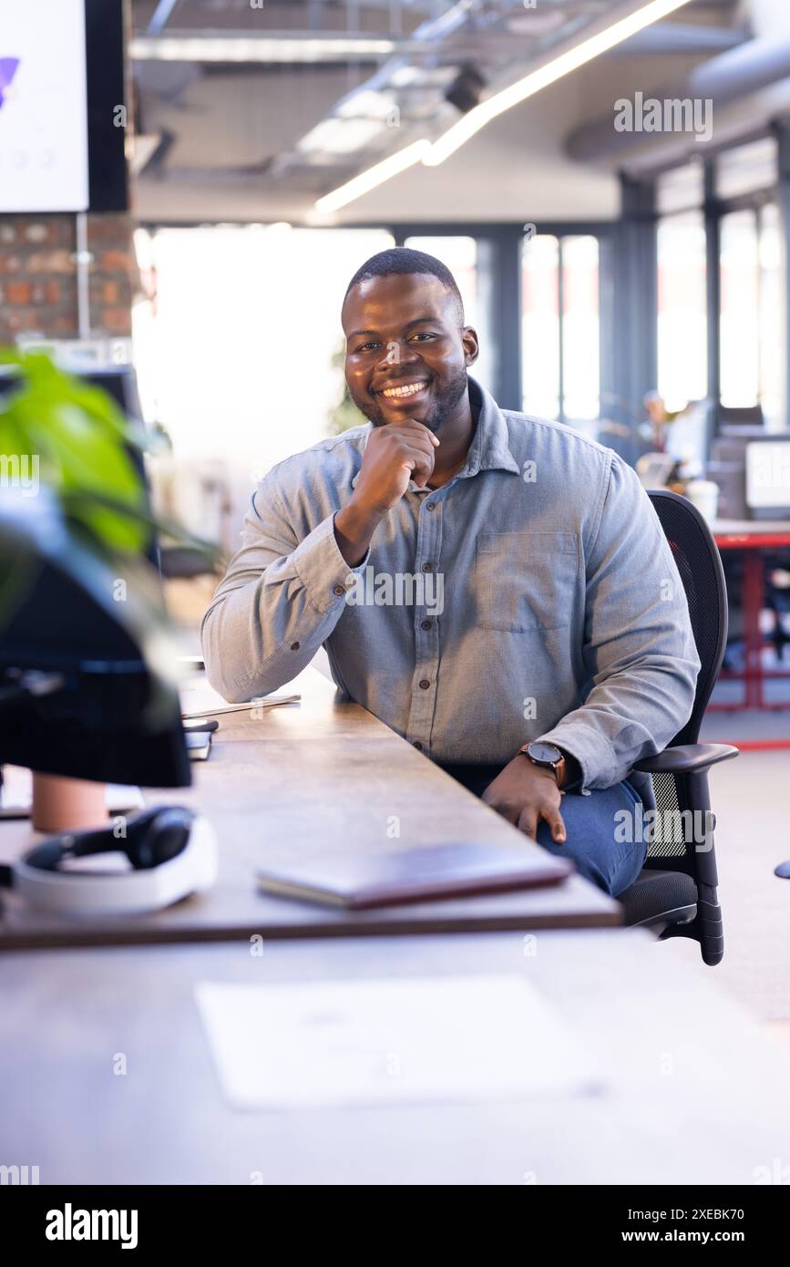 Lächelnder afroamerikanischer Geschäftsmann, der am Schreibtisch in einem modernen Büroarbeitsplatz sitzt Stockfoto