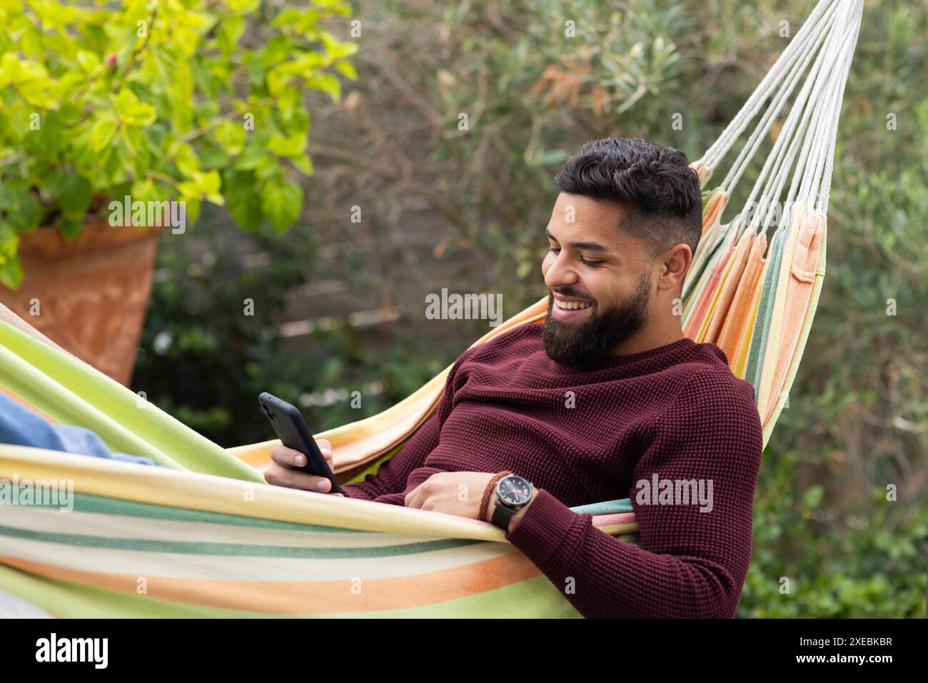 Lächelnd und mit dem Smartphone, Mann entspannt sich in der Hängematte draußen Stockfoto