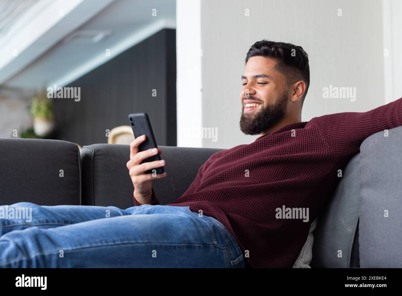 Entspannen Sie sich auf der Couch, ein Mann lächelt, während er zu Hause das Smartphone benutzt Stockfoto