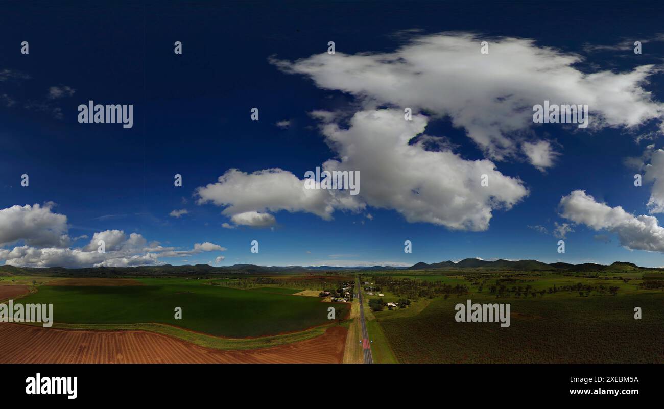 Panoramablick mit blauem Himmel, flauschigen Wolken über grünen Feldern und einer langen geraden Straße Stockfoto