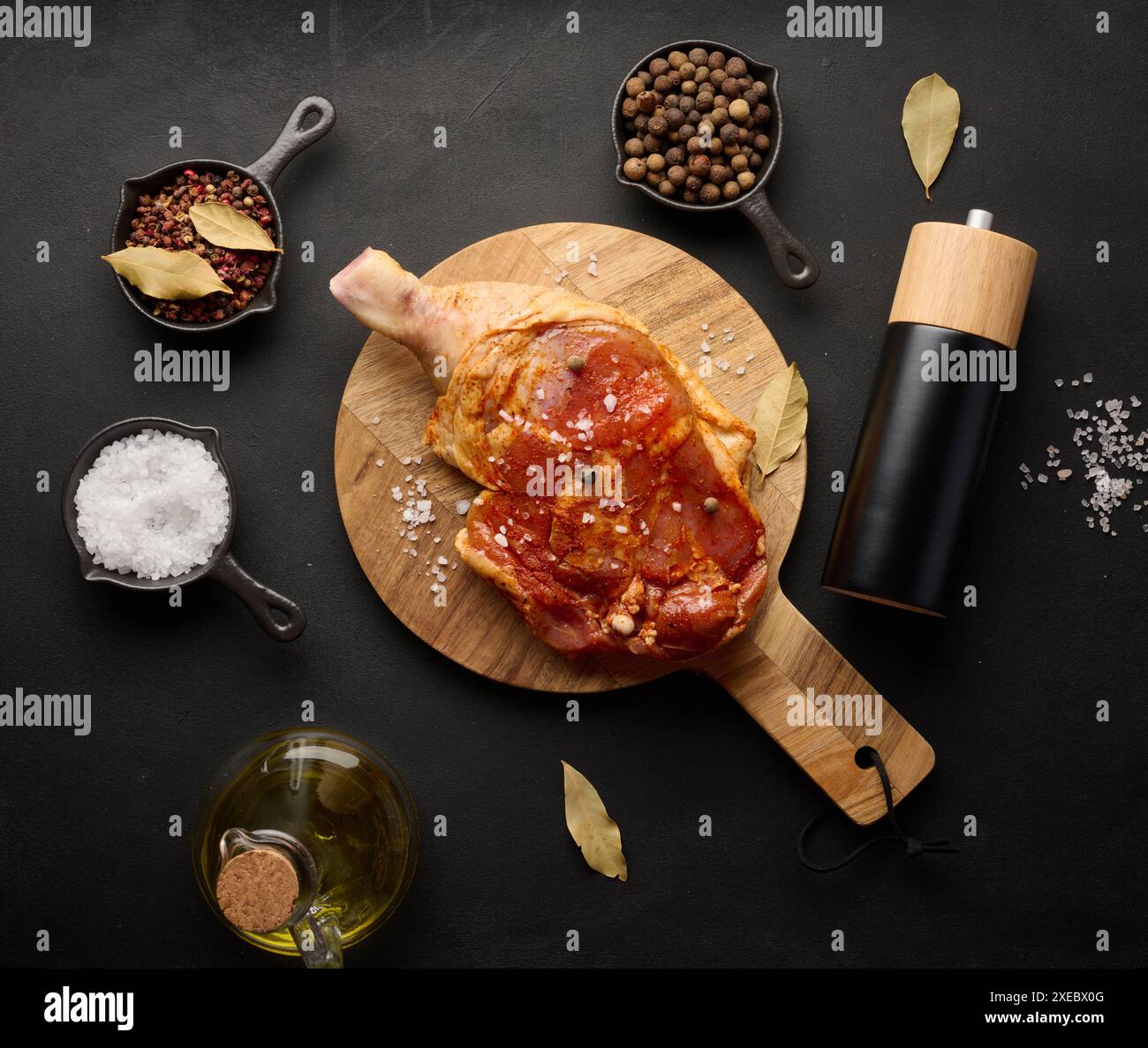 Rohe Hähnchenkeulen, gewürzt auf einem Holzbrett, begleitet von Salz und Pfeffer, von oben gesehen. Schwarzer Tisch Stockfoto