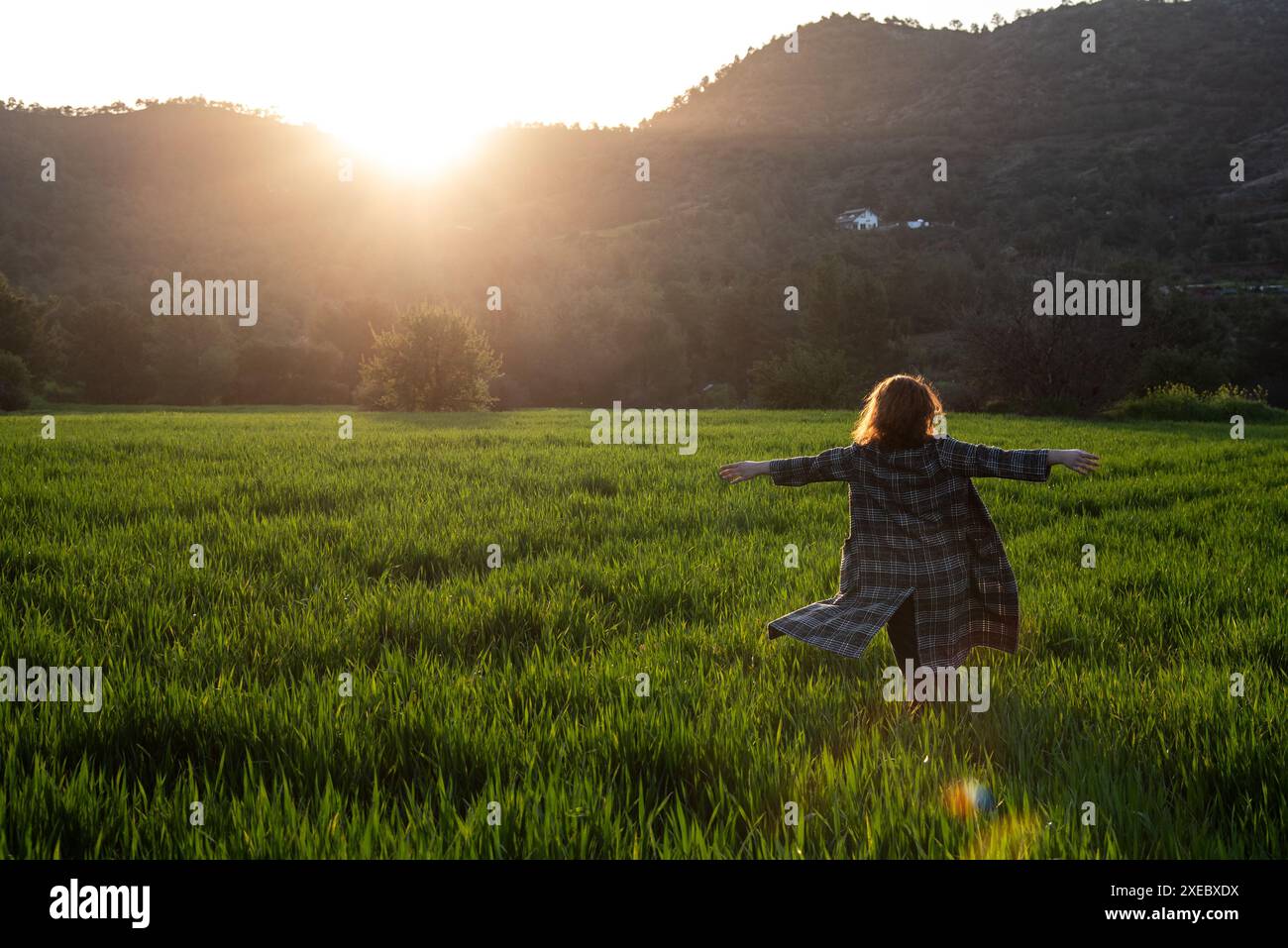 Frau mit langem Fell und gestreckten Armen genießt Sonnenuntergang auf der grünen Wiese Stockfoto