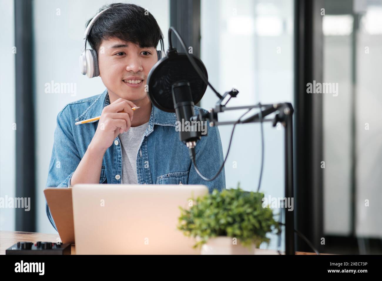 Ein Mann mit Kopfhörern und blauem Hemd sitzt mit einem Laptop an einem Schreibtisch Stockfoto