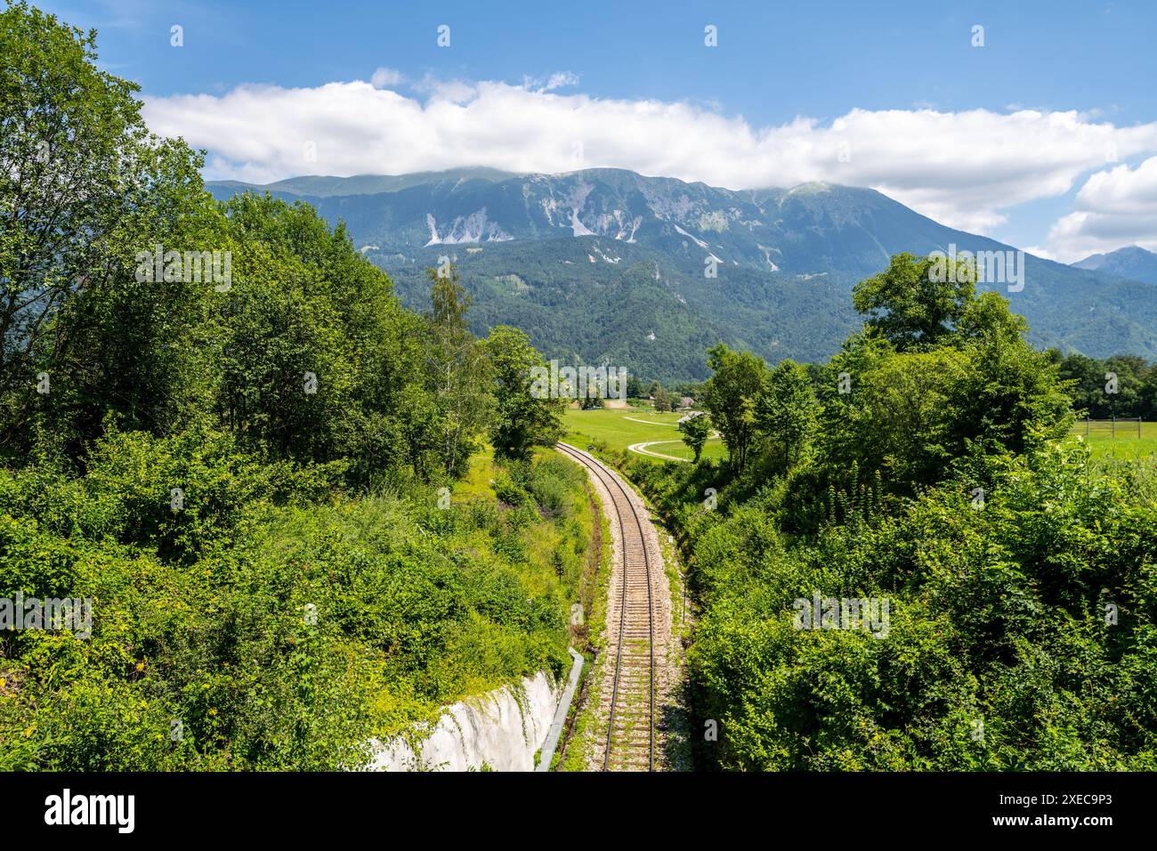 Eine Eisenbahn schlängelt sich durch ein üppiges Tal mit den Julischen Alpen im Hintergrund. Genießen Sie eine malerische Zugfahrt durch die atemberaubende Landschaft Sloweniens an einem sonnigen Sommertag. Stockfoto