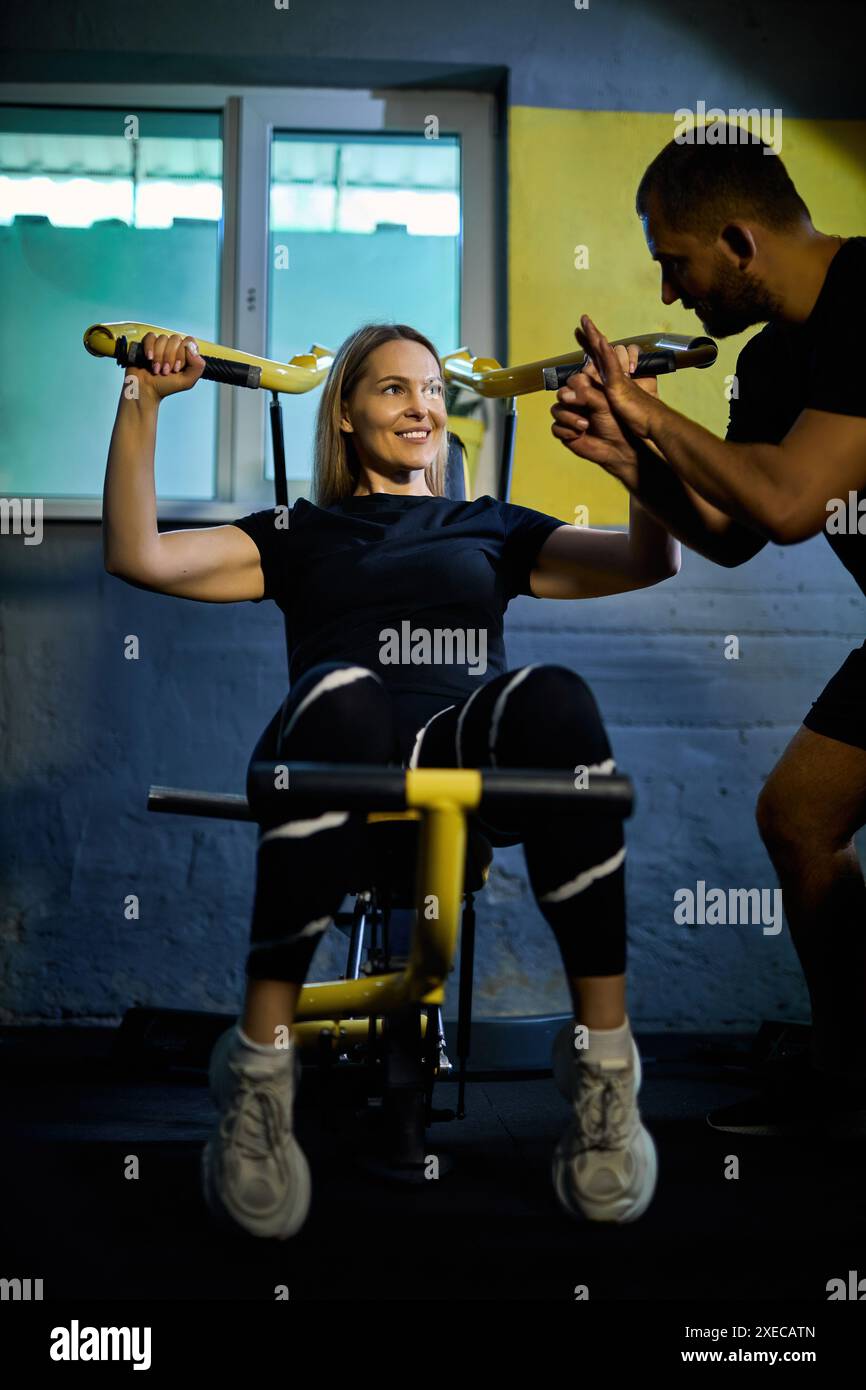 Personal Coach, der die Sportlerin bei der Benutzung des Geräts während des Fitnessstudios in einem dunklen Fitnessstudio unterstützt Stockfoto