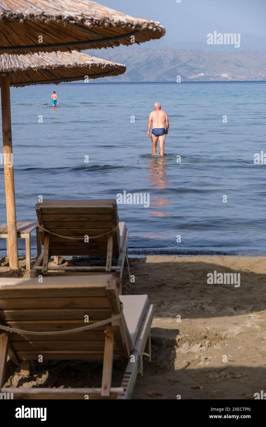 Großer Schilfschirm an einem Strand auf Korfu in Griechenland, mit ruhigem tiefblauem Meer und Liegestühlen am Sandstrand. Stockfoto