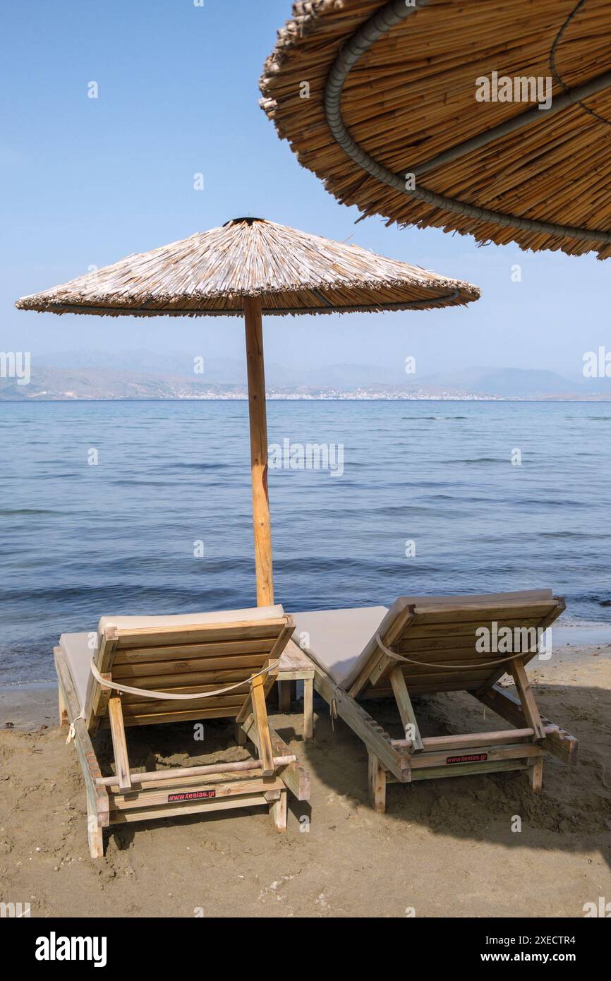 Großer Schilfschirm an einem Strand auf Korfu in Griechenland, mit ruhigem tiefblauem Meer und Liegestühlen am Sandstrand. Stockfoto