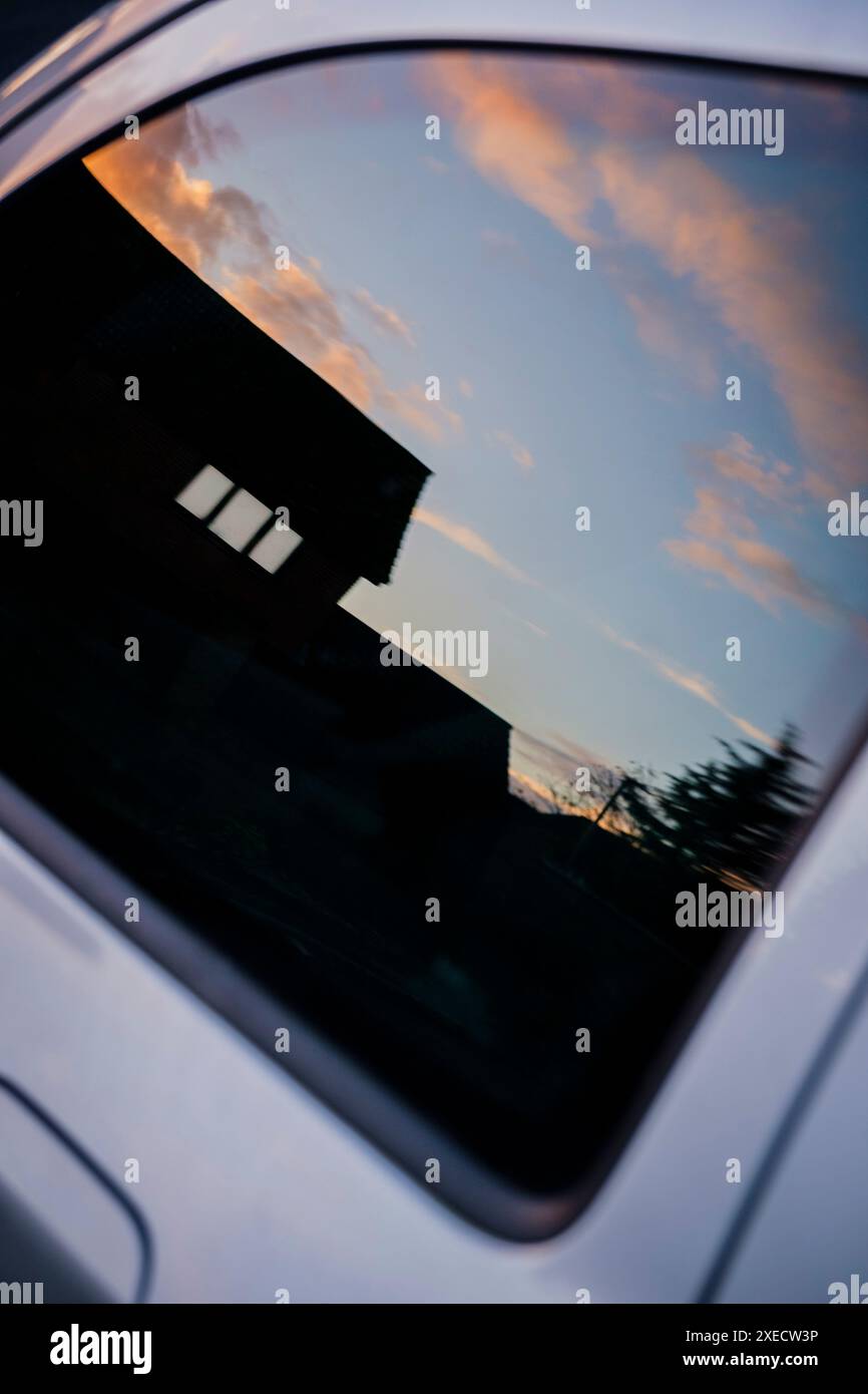 Reflexion eines Hauses in Silhouette in einem Seitenfenster des Autos mit einigen Häuserfenstern und einem Abendhimmel. Stockfoto