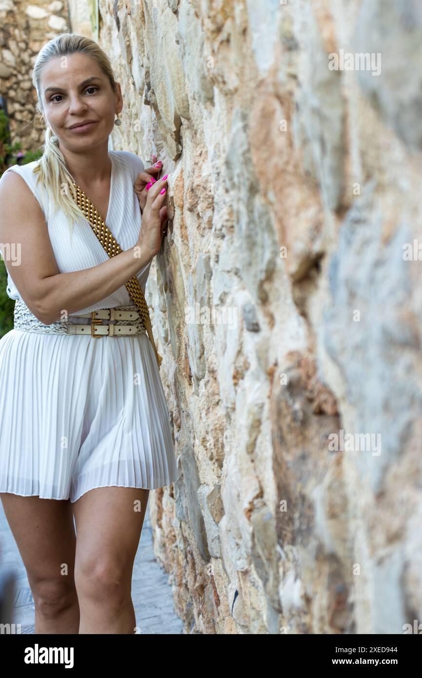 Dieses Bild zeigt eine schöne blonde Frau, die sich gegen eine Steinmauer posiert. Sie ist elegant in einem weißen Kleid gekleidet und mit einem goldenen Gürtel versehen Stockfoto