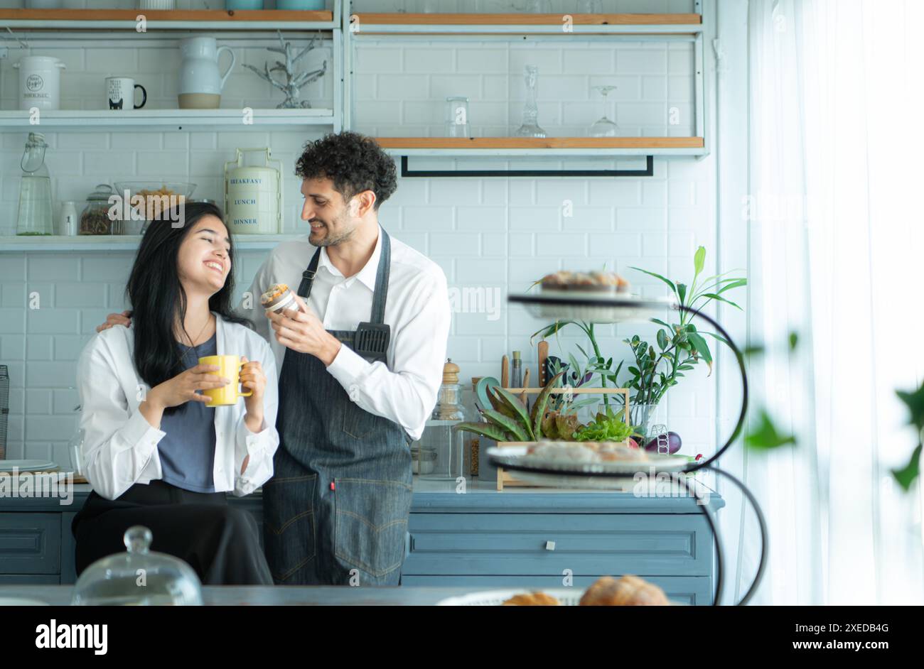 Ein Ehemann trägt eine Schürze, während er mit seiner Frau ein Abendessen kocht. Das Duo machte eine Pause, um Kaffee zu trinken und das fertige Brot zu probieren. Stockfoto